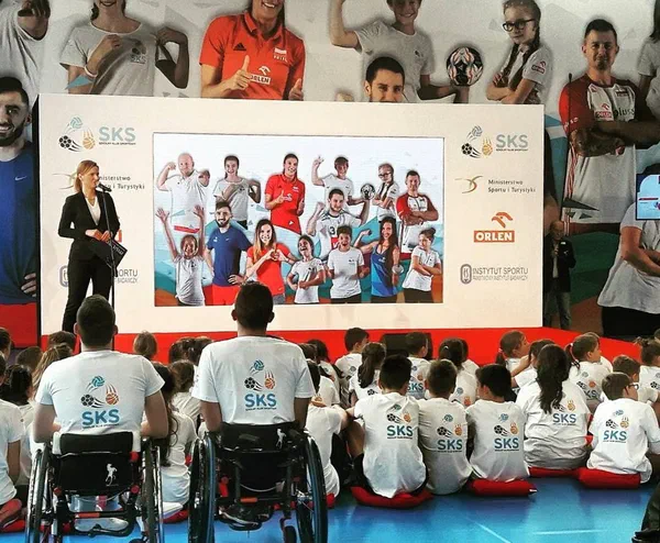 Konferencja programu Szkolny Klub Sportowy w hali sportowej: dzieci i młodzież w koszulkach SKS (w tym zawodnicy na wózkach sportowych) przed sceną z ekranem i sportowcami.