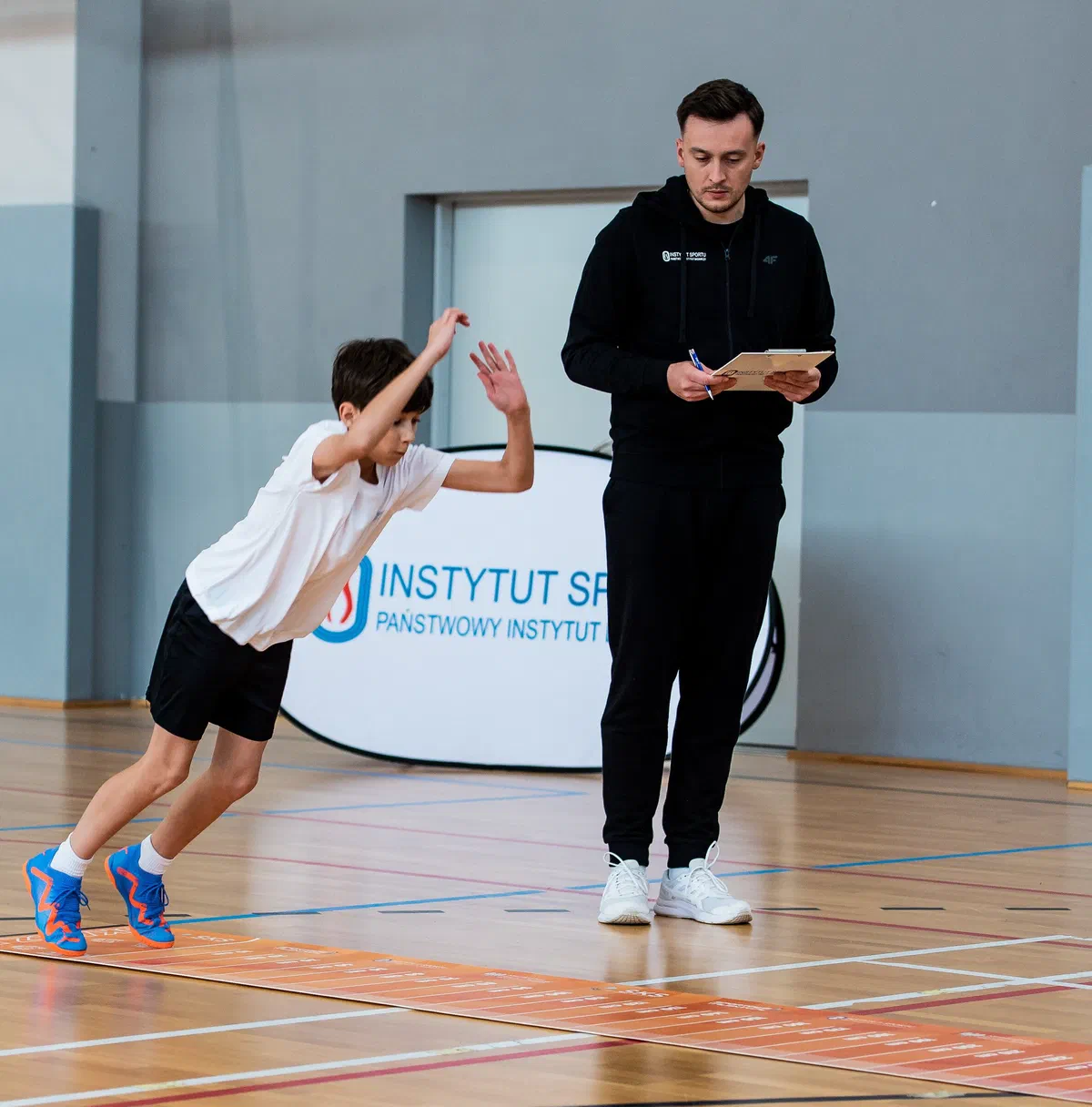 Uczeń wykonujący skok w dal z miejsca na macie pomiarowej w sali gimnastycznej pod okiem nauczyciela podczas testów programu Sportowe Talenty.