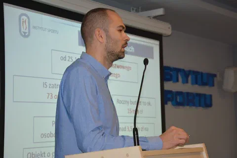 Prelegent w jasnoniebieskiej koszuli przemawiający przy mównicy na tle ekranu ze schematem organizacyjnym