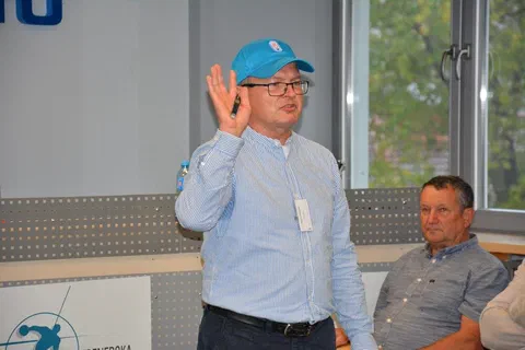 Prelegent w jasnej koszuli i niebieskiej czapce przemawiający z podniesioną ręką przed napisem Instytut Sportu