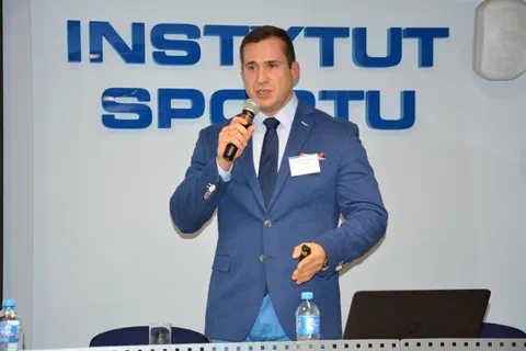 Prelegent w niebieskim garniturze i krawacie przemawiający do mikrofonu na tle napisu Instytut Sportu