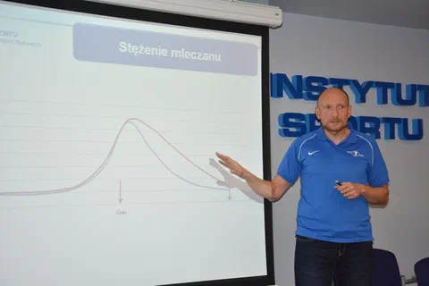 Wykładowca w niebieskiej koszulce polo wskazujący na ekranie wykres zmian stężenia mleczanu