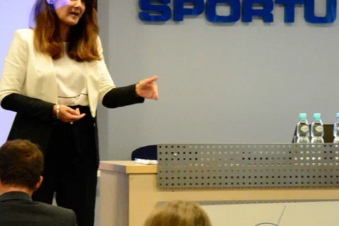 Prelegentka w białym żakiecie występująca przed stołem prezydialnym i napisem Instytut Sportu