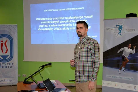 Wykładowca omawiający slajd o kształtowaniu otoczenia wspierającego rozwój utalentowanych sportowców obok baneru Akademii Trenerskiej