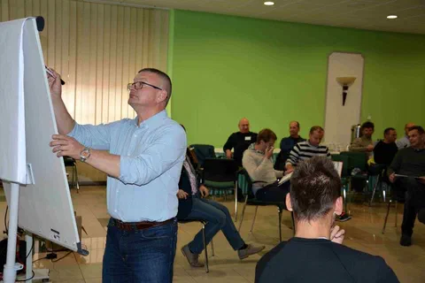 Prowadzący piszący na flipcharcie przed uczestnikami siedzącymi w grupach w głębi sali