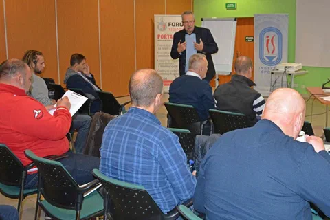 Uczestnicy siedzący w podgrupach i słuchający prowadzącego stojącego z przodu sali na tle banerów