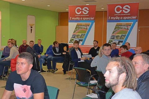 Trenerzy siedzący w półkolu pod czerwonymi banerami Centralnego Ośrodka Sportu podczas zajęć warsztatowych