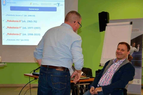 Prowadzący i siedzący obok uczestnik przed ekranem ze slajdem o pokoleniach X, Y, Z i Alfa