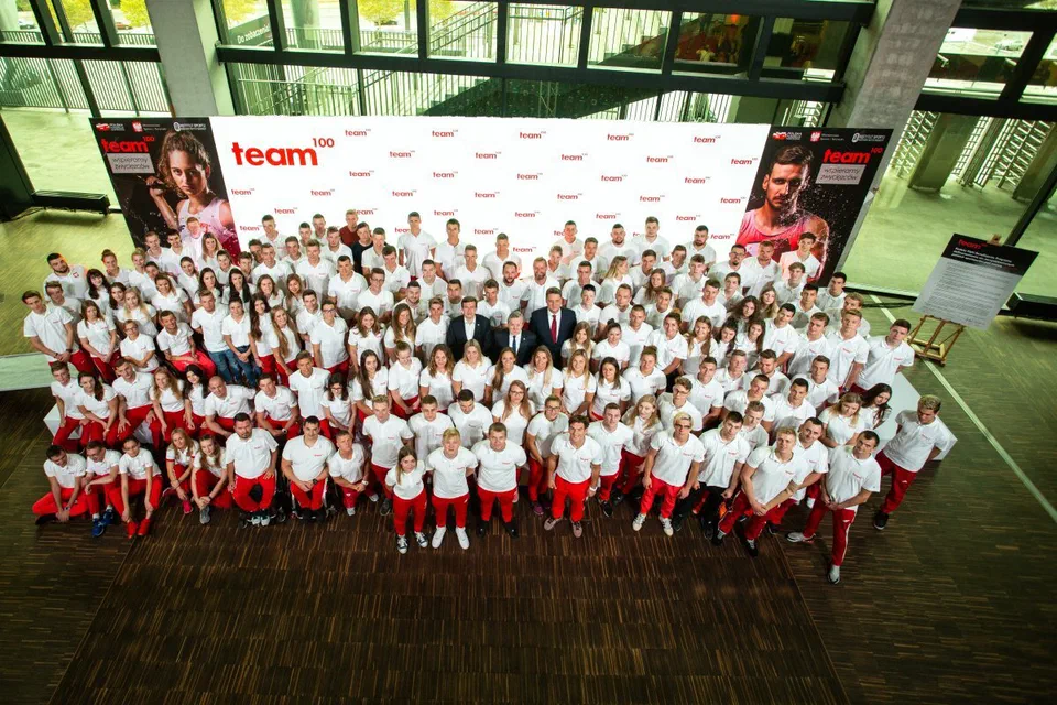 Grupowe zdjęcie beneficjentów programu Team100 w biało-czerwonych strojach wraz z przedstawicielami rządu, Polskiej Fundacji Narodowej i Instytutu Sportu – PIB na Stadionie Narodowym