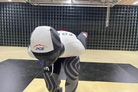 Łyżwiarz szybki w kasku i kombinezonie reprezentacji Polski ustawiony w niskiej pozycji w tunelu aerodynamicznym