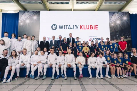 Wspólne zdjęcie młodych sportowców w strojach szermierczych i piłkarskich z gośćmi na tle ekranu z hasłem Witaj w Klubie