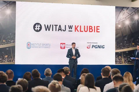 Wystąpienie na scenie przed dużym ekranem z hasłem Witaj w Klubie i logotypami Instytutu Sportu, ministerstwa oraz sponsora