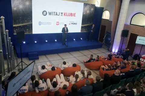 Widok z balkonu na salę konferencyjną z uczestnikami siedzącymi na pomarańczowych pufach i krzesłach przed sceną