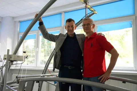 Dwaj mężczyźni stojący na pomoście przy dużej bieżni pomiarowej z uprzężą zabezpieczającą w laboratorium