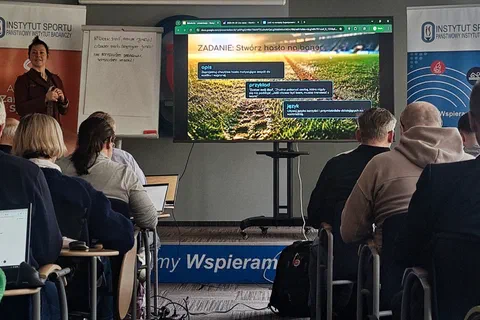 Prowadzący omawiający zadanie zespołowe wyświetlone na ekranie przed uczestnikami pracującymi przy laptopach