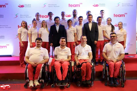 Grupa zawodników programu Team100, w tym sportowców na wózkach, pozująca z przedstawicielami instytucji na tle ścianki programu