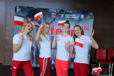 Czworo młodych sportowców w biało-czerwonych strojach pozujących z chorągiewkami na tle baneru Team100