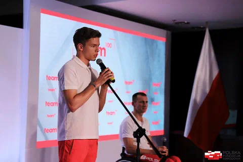 Młody zawodnik przemawiający do mikrofonu na scenie obok zawodnika na wózku i flagi państwowej