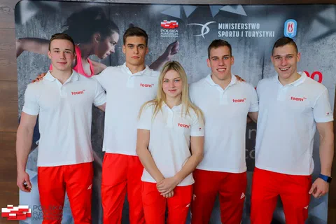 Pięcioro beneficjentów programu Team100 w białych koszulkach i czerwonych spodniach dresowych pozujących na tle plakatu
