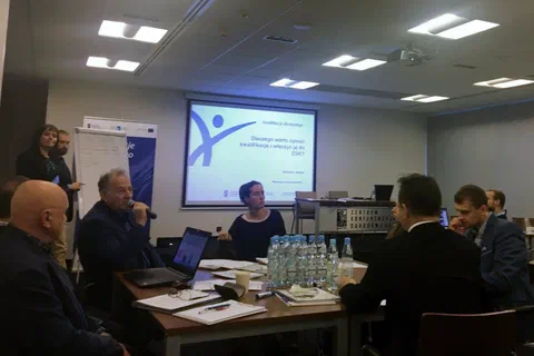 Uczestnicy spotkania roboczego dyskutujący przy stole z mikrofonem i laptopami na tle ekranu z logo Akademii Trenerskiej