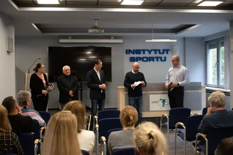 Pięciu mężczyzn stojących z przodu sali i przemawiających do uczestników wernisażu wystawy