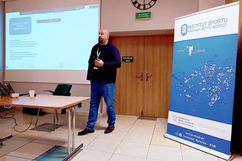 Wykładowca przemawiający przez mikrofon obok ekranu projekcyjnego i niebieskiego roll-upu Instytutu Sportu