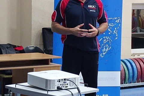 Prelegent w stroju sportowym przemawiający na tle roll-upu przed projektorem