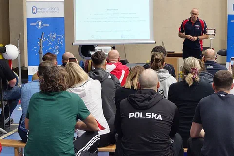 Wykładowca w stroju sportowym omawiający schemat na ekranie przed uczestnikami siedzącymi w sali