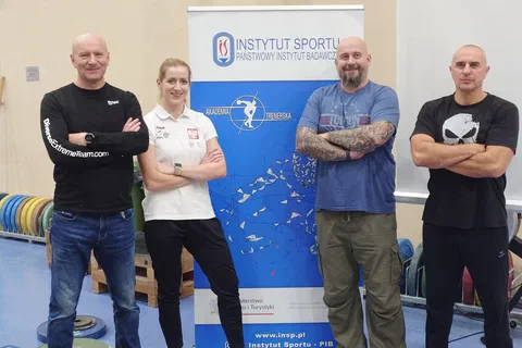Czworo prowadzących w strojach sportowych pozujących obok roll-upu Instytutu Sportu w sali ze sprzętem ciężarowym