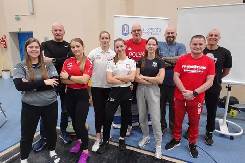Grupa ośmiorga uczestników i prowadzących w strojach treningowych pozująca w sali ćwiczeń obok sztangi i tablicy