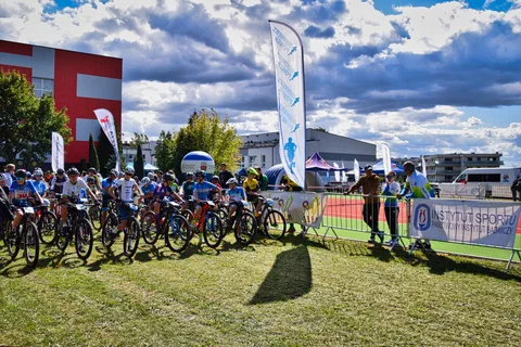 Młodzi kolarze górscy ustawieni w rzędzie na linii startu na trawiastej polanie obok banerów Instytutu Sportu