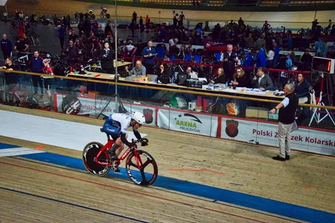 Pojedynczy kolarz w biało-niebieskim stroju jadący w niskiej pozycji aerodynamicznej wzdłuż sędziów