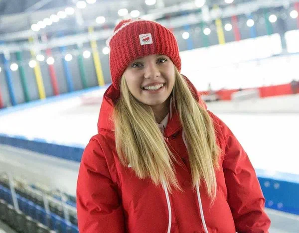 Panczenistka Karolina Bosiek w czerwonej kurtce i czapce reprezentacyjnej na tle krytego toru łyżwiarskiego.