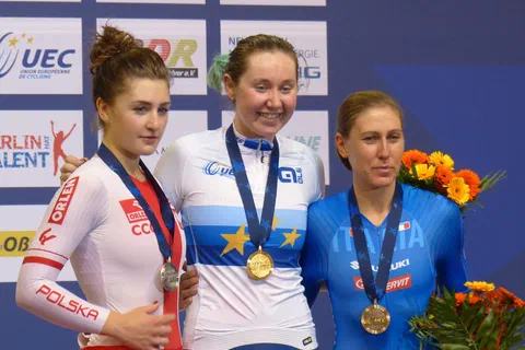 Trzy kolarki stojące na podium mistrzostw Europy z medalami na szyjach i bukietami kwiatów