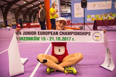 Zwyciężczyni siedząca za linią mety na podłodze hali i unosząca nad głową szarfę z nazwą zawodów w Drzonkowie